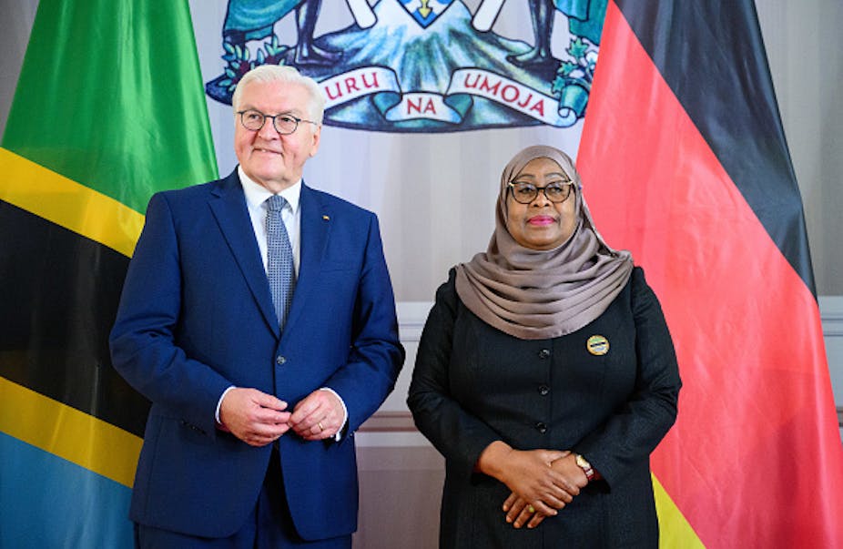 A man with white hair, wearing a suit, stands in front of flags alongside a woman wearing a headscarf.