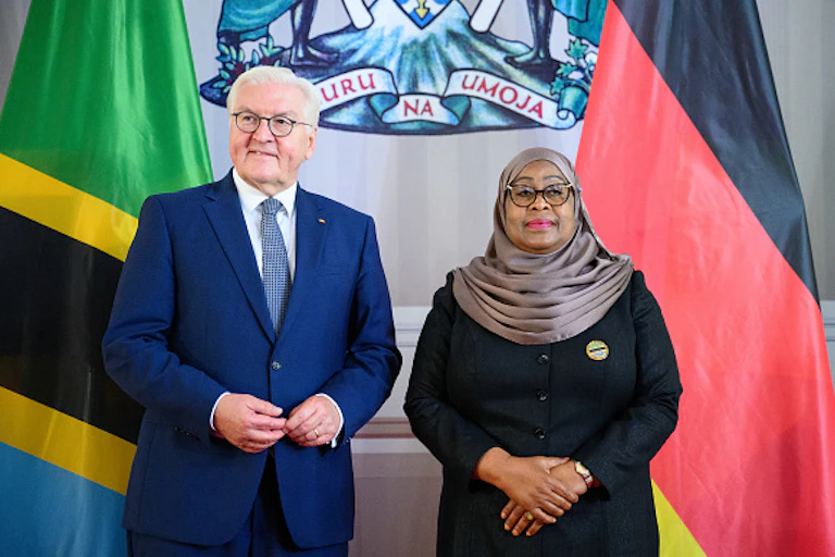 A man with white hair, wearing a suit, stands in front of flags alongside a woman wearing a headscarf.