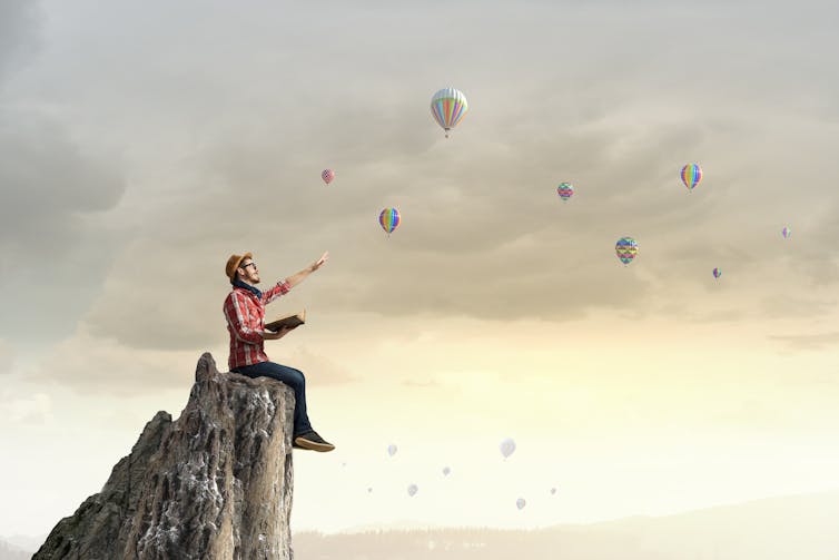 Photo montage of a man sitting on a mountain with a book, facing hot-air balloons.