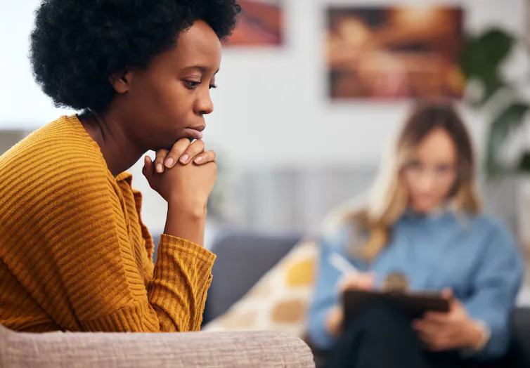 A young woman sits sadly on a couch with her head on her hands, a therapist sits on a couch and takes notes in the background