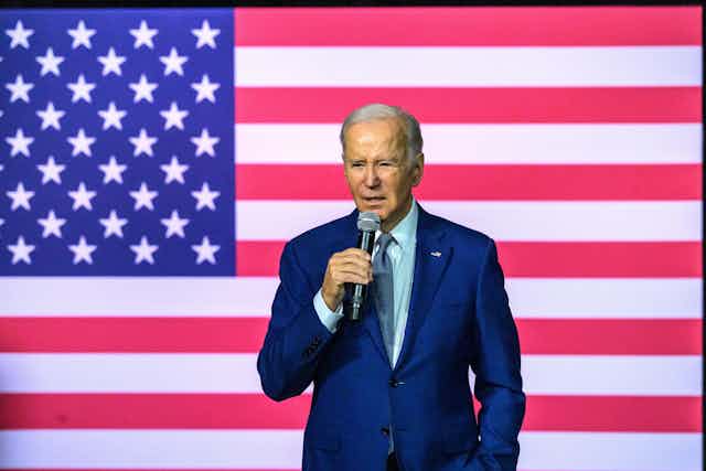 US president Joe Biden holds a microphone standing in front of a US flag.
