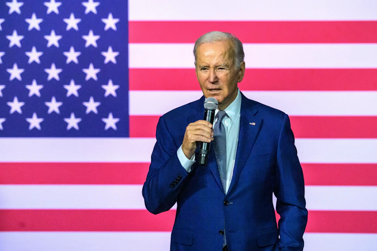 US president Joe Biden holds a microphone standing in front of a US flag.