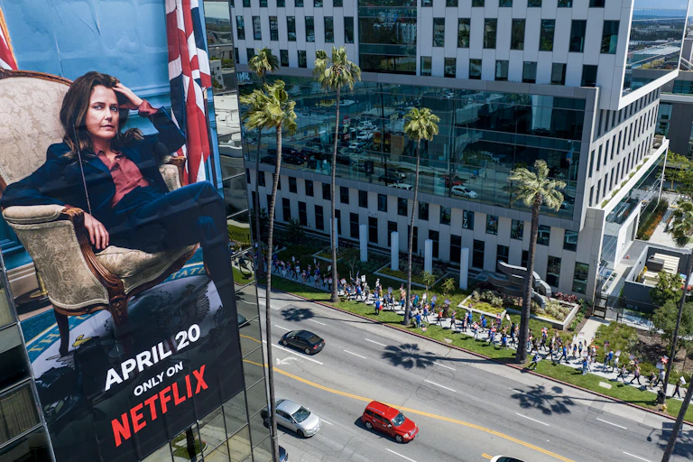 The actress Keri Russell sits in a chair in a billboard for the show 'The Diplomat' hung on a building above people walking as part of a protest on a Los Angeles street.