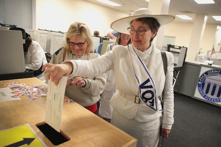 Women in white drop a ballot in a voting box.