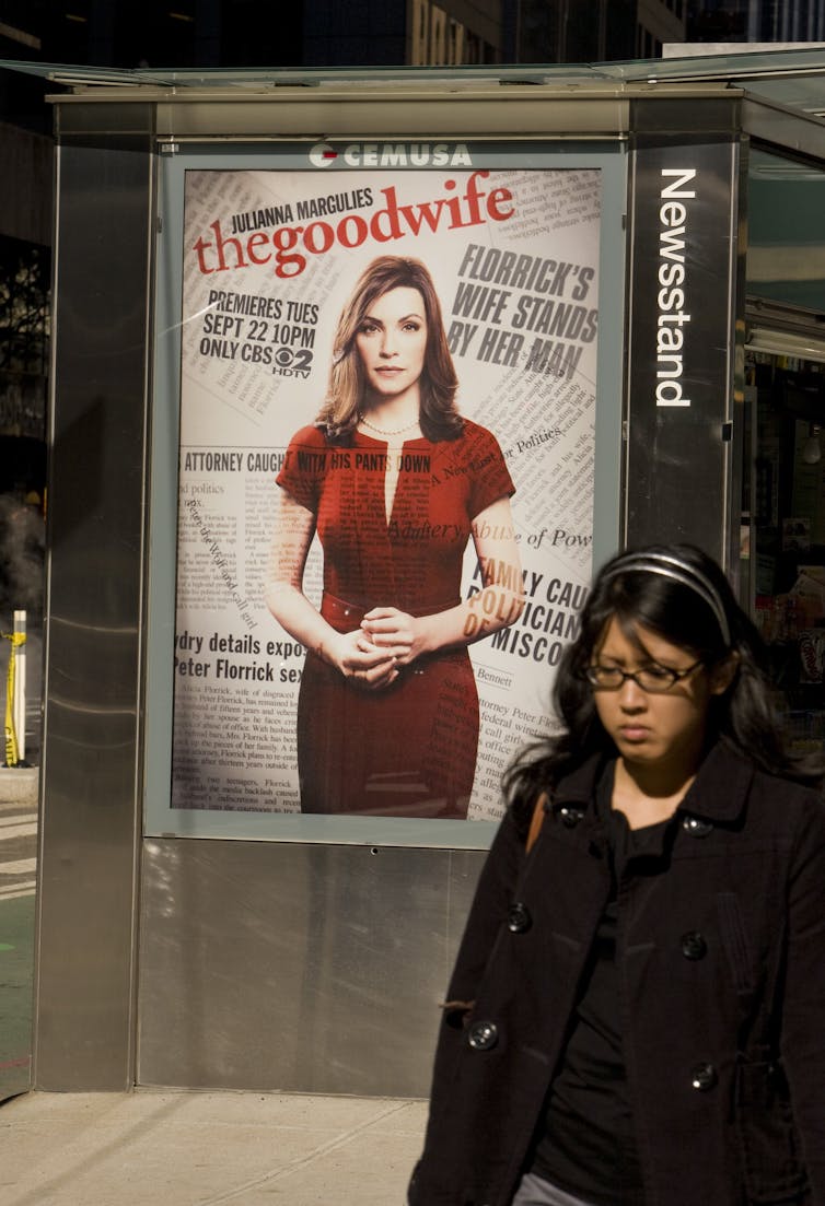 A woman walks past a billboard in front of a bus stop in a city. The billboard is for the show 'The Good Wife,' and shows a middle aged woman in a red dress looking directly at a camera.