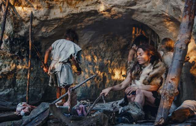 people dressed in skins outside a cave, a woman seated at a fire
