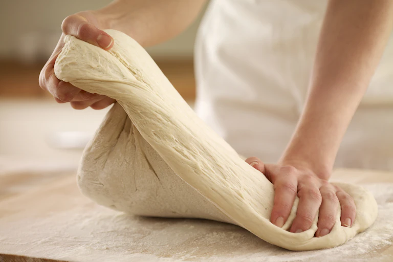 A person's hands holding and stretching a light brown dough, dusted with flour. They're wearing a white apron and the brown table they're working at is dusted with flour.
