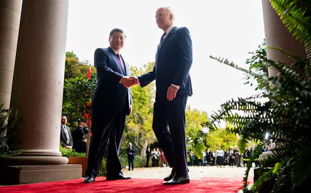Two men shake hands while standing on a red carpet.