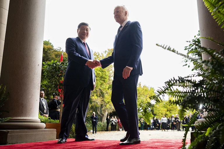 Two men shake hands while standing on a red carpet.