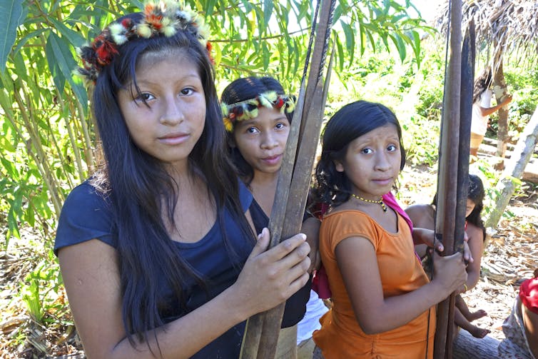 young women adorned with toucan and macaw feathers holding wooden sticks