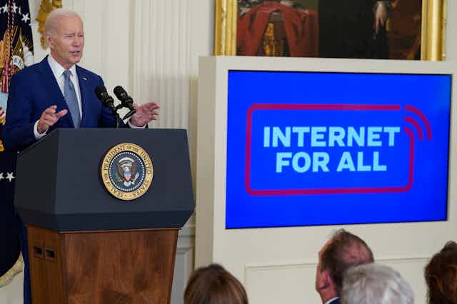 An older man in a business suit stands at a podium with a circular emblem on the front