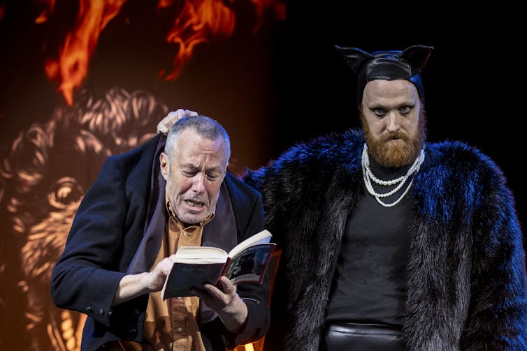 A man reads a book, another man dressed as a cat holds him by the nape of his neck.