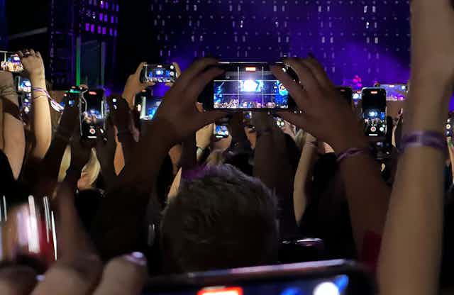 A photo taken in the audience at a concert, showing crowd members holding up their phones to photograph the stage.