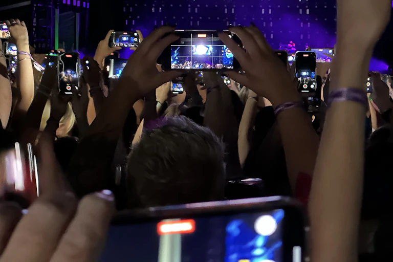 A photo taken in the audience at a concert, showing crowd members holding up their phones to photograph the stage.