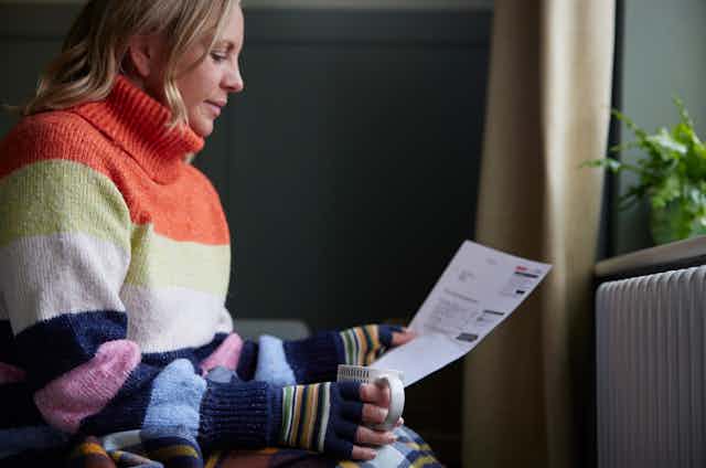 Woman in jumper and gloves looking at a bill beside a radiator.