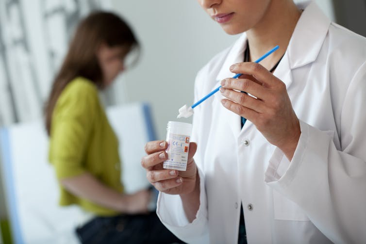 A doctor places a smear test swab in a vial while a female patients sits in the background on an examination table.