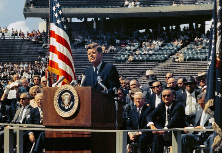 JFK stands by a US flag in a stadium.