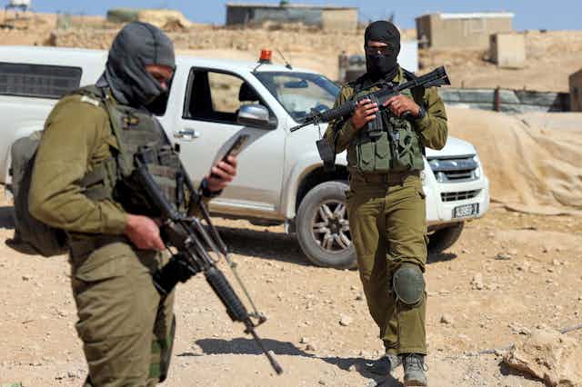 Two soldiers with guns and face masks standing on rocky, sandy soil.