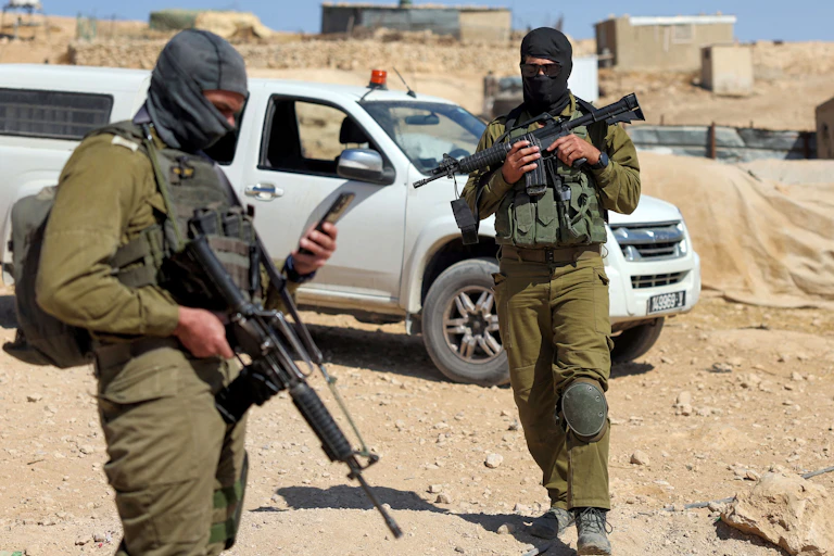 Two soldiers with guns and face masks standing on rocky, sandy soil.
