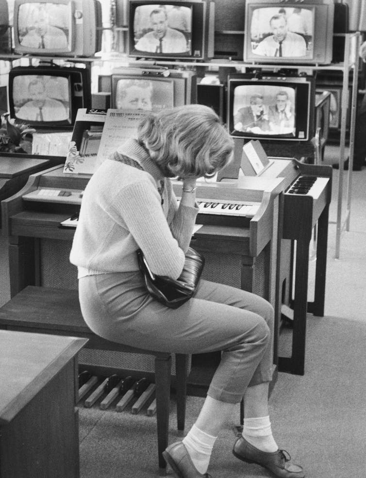 In a black and white image, a young woman is seen crying in front of half a dozen televisions.