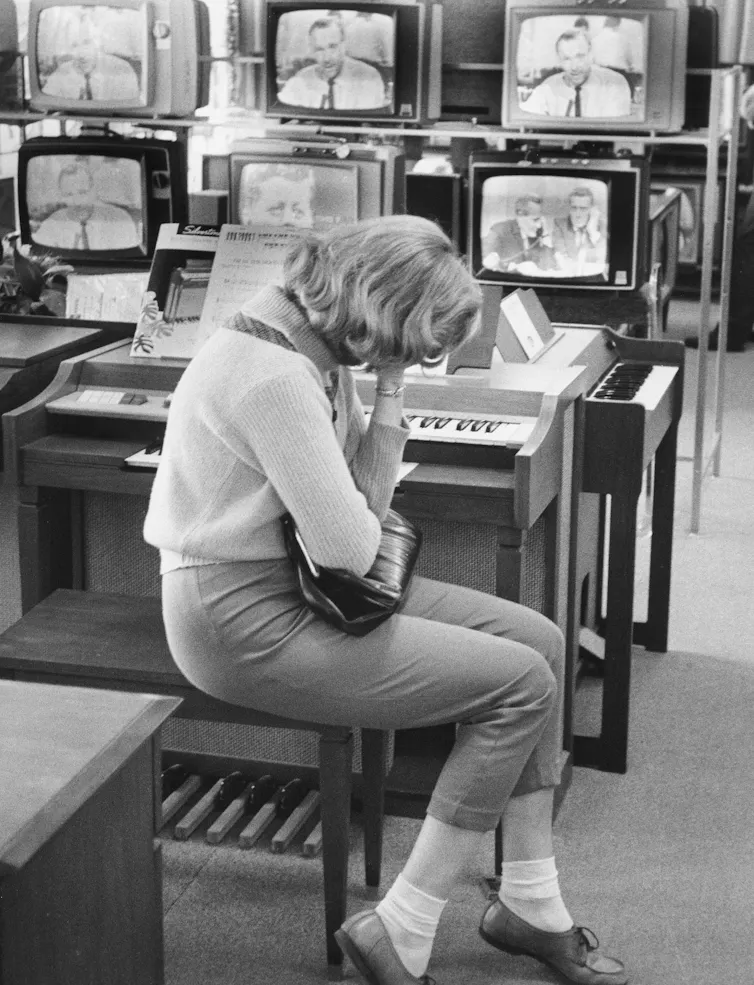 In a black and white image, a young woman is seen crying in front of half a dozen televisions.