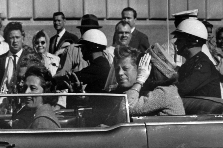 A black and white photo shows JFK and Jacqueline Kennedy in a convertible car. Onlookers are visible in the background.