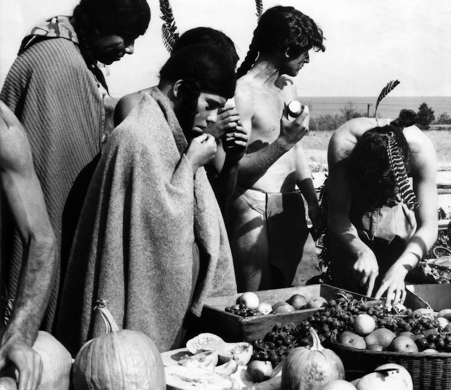 A black and white picture of men and women in traditional Native American dress seen feasting on food in front of them.