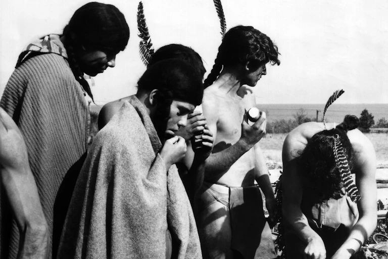 A black and white picture of men and women in traditional Native American dress seen feasting on food in front of them.