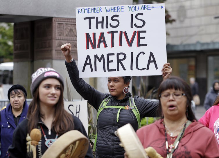 A woman protestor, standing with others, holding a sign that says 'This is Native America.'