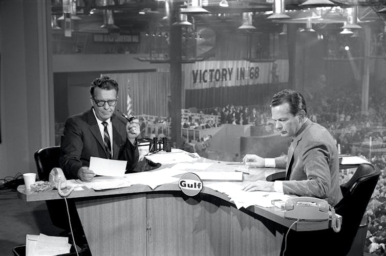 In a black-and-white photo, two news anchors, one smoking a pipe, are seen sitting in a broadcast studio at the Miami Beach Convention Center. In the background, conventioneers are seen milling around and a sign reads 'VICTORY IN 68'