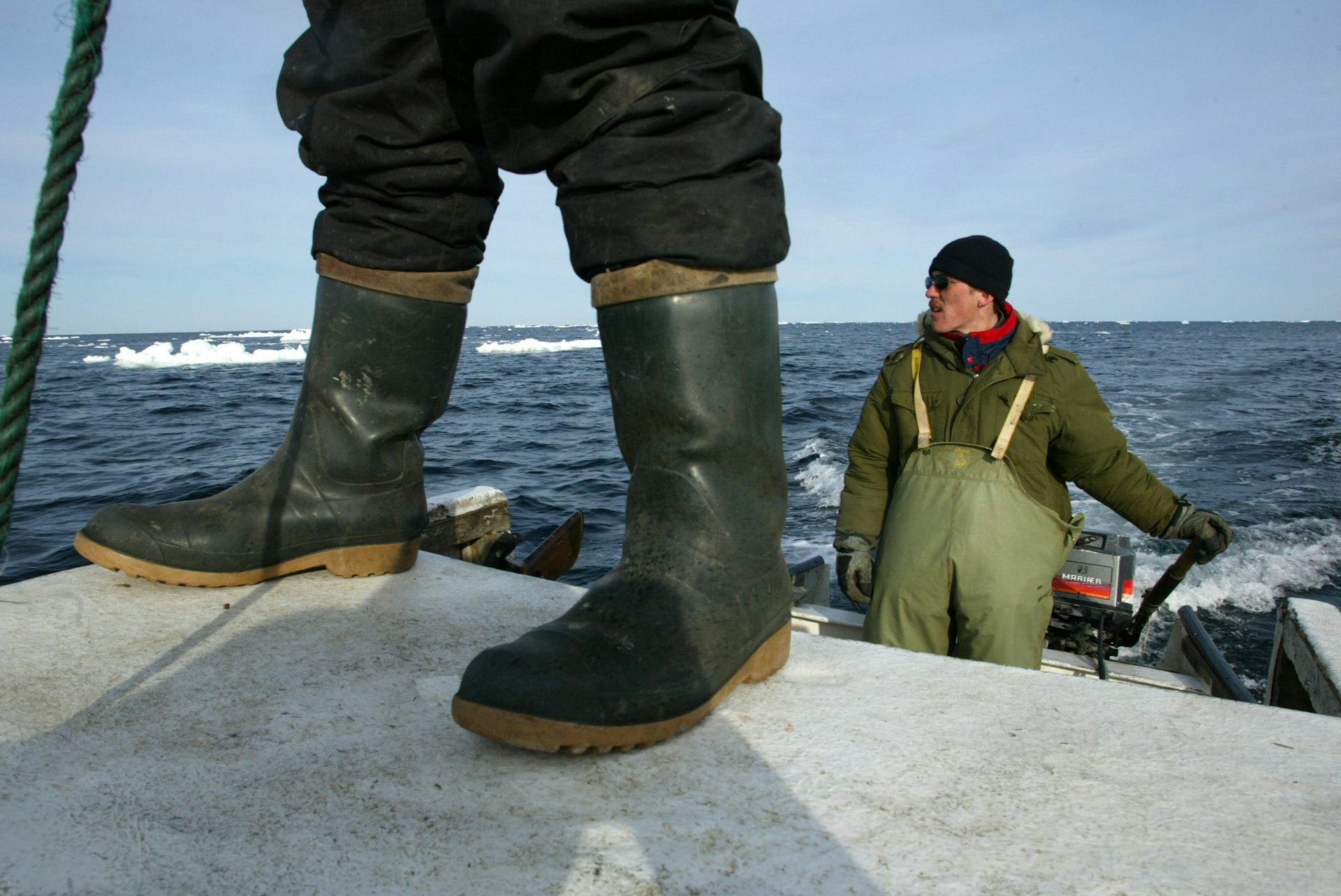 The rubber boots of a man are seen in front of another man who steers a boat in icy water.