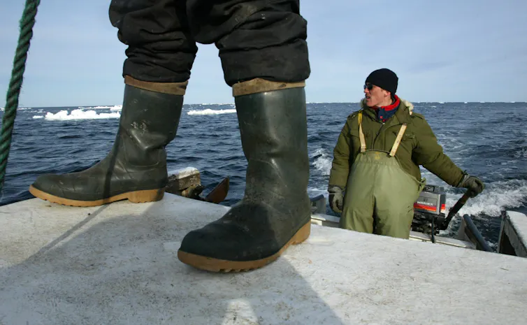 The rubber boots of a man are seen in front of another man who steers a boat in icy water.