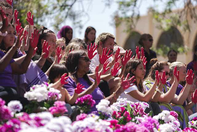 A group of people sit among flowers with red on their hands as part of a Day of Action. This is in response to the lack of funding and support for domestic and family violence support services in the Northern Territory.