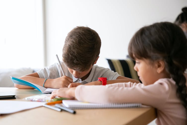 Two young children work at a desk with books and pens.