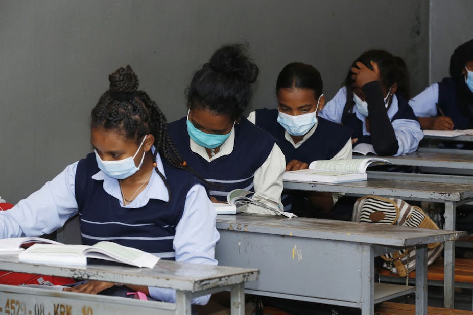A photograph of five learners, each sitting at desks one behind the other. They are all wearing masks and studying their books