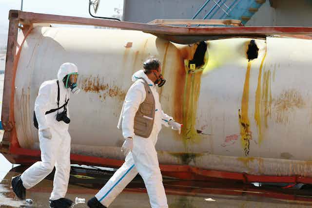 Two men in hazmat suits, wearing respirators, walk past a large ruptured metal tank