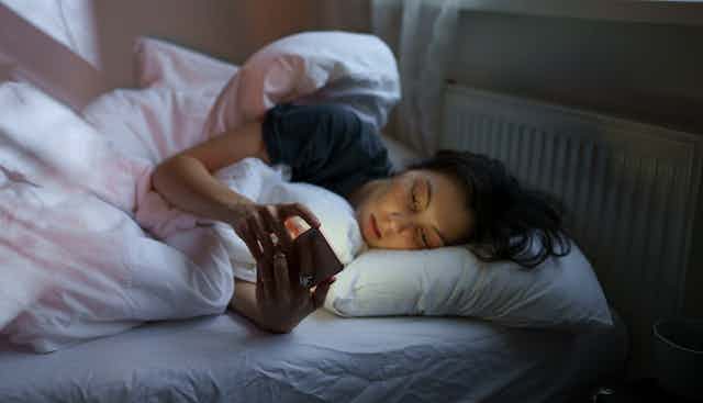 A young woman lying in bed scrolling on her phone