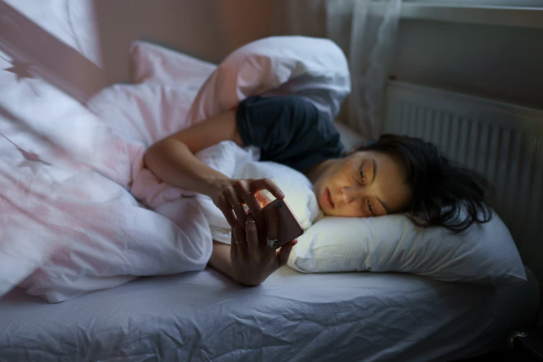 A young woman lying in bed scrolling on her phone