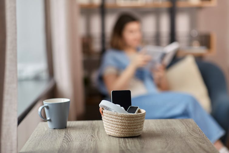 A smartphone sitting in a small basket on top of a table, in the background a woman is sitting on a chair reading