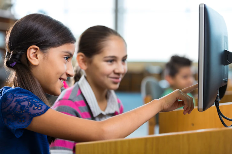 Two smiling girls are seated next to each other as they look at a computer screen.