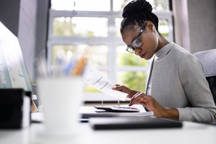 Woman with glasses at a desk by a window, calculator, paper work, computer.