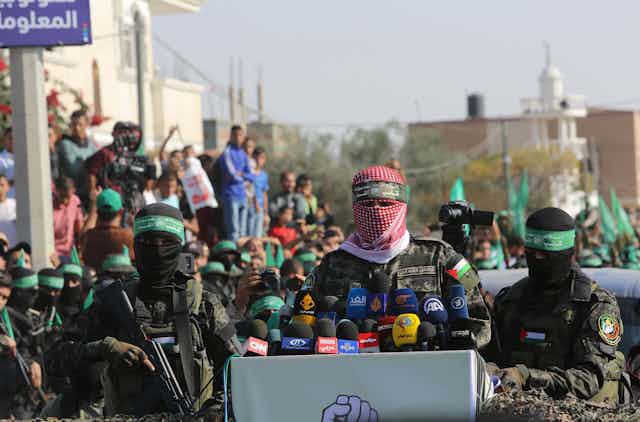 Des hommes masqués aux couleurs du Hamas prononcent un discours devant des micros. Derrière eux, des civils sont rassemblés.