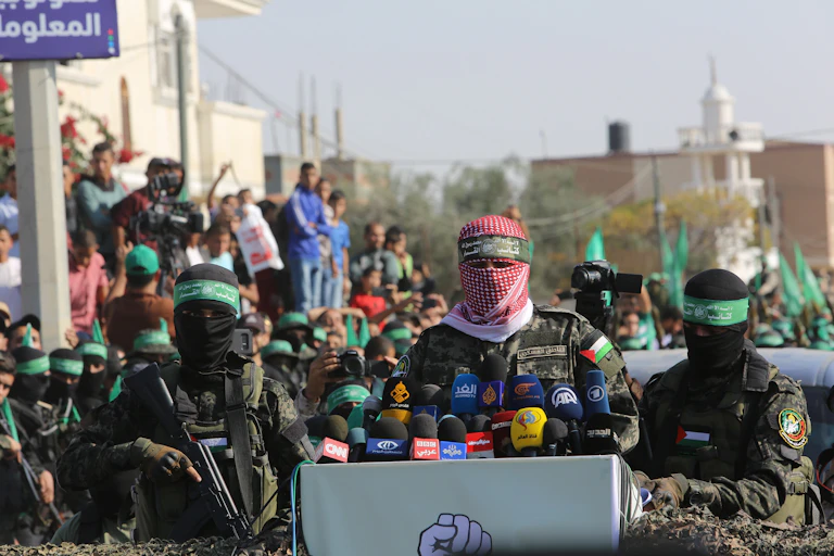 Des hommes masqués aux couleurs du Hamas prononcent un discours devant des micros. Derrière eux, des civils sont rassemblés.