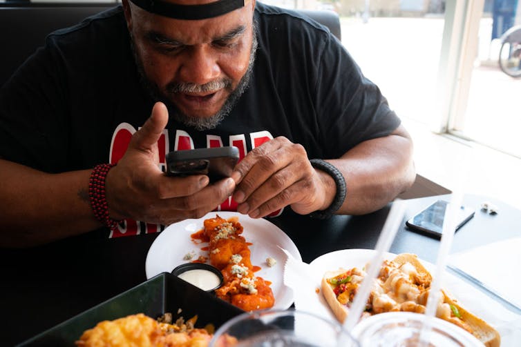 Large Black man hunches over a meal of fried seafood as he holds his smartphone.