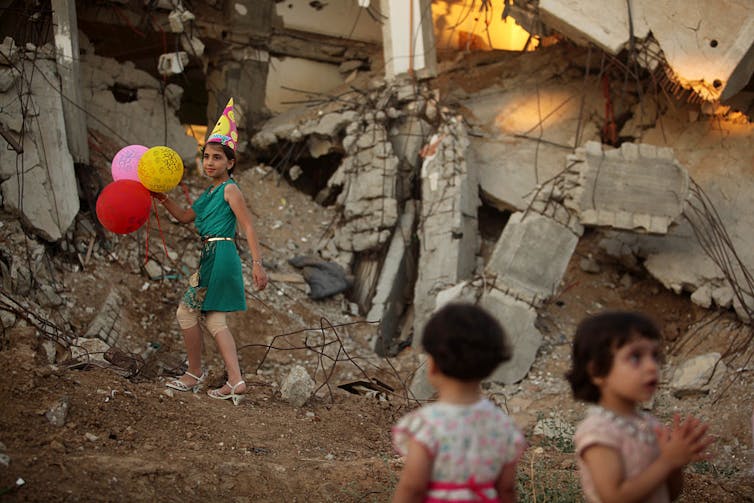 A girl wears a birthday hat and holds three balloons in front of a destroyed building.
