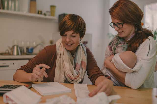 A woman holding a baby chats with her female partner about household expenses.