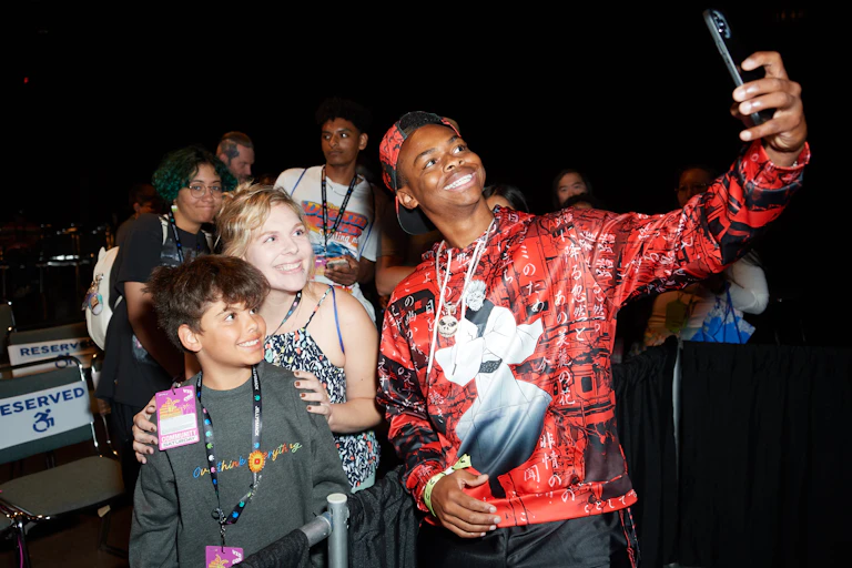Black teenager wearing red shirt and red hat takes a selfie with smiling fans.
