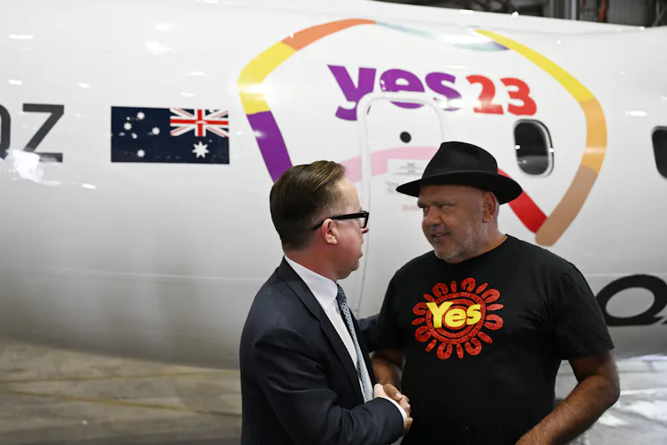 Two men standing and talking with a plane in the background.