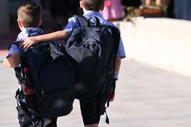 Two primary school students walk with backpacks. One student has an arm around the other.