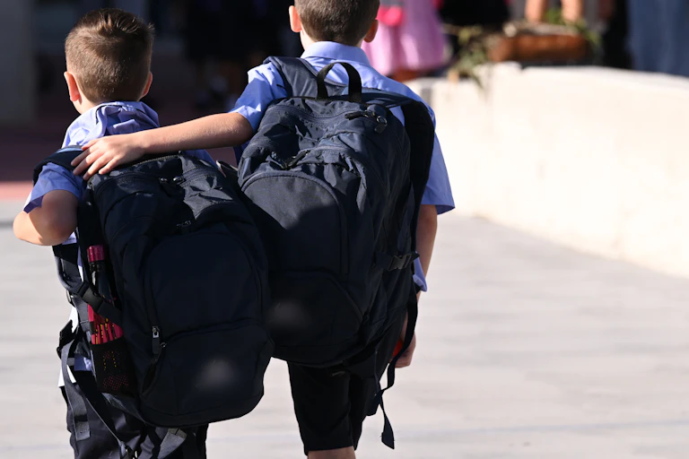 Two primary school students walk with backpacks. One student has an arm around the other.